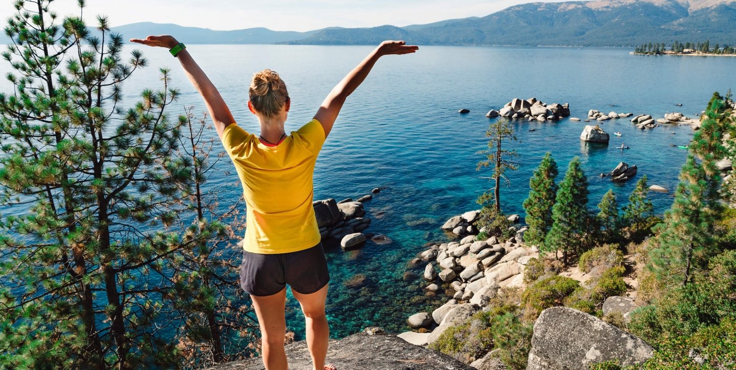 A backshot of a woman with her arms raised next to the blue water and mountains
