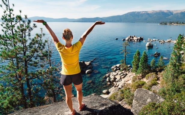 A backshot of a woman with her arms raised next to the blue water and mountains