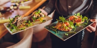 A server's hands carrying two plates of open-faced sandwiches garnished with mint.