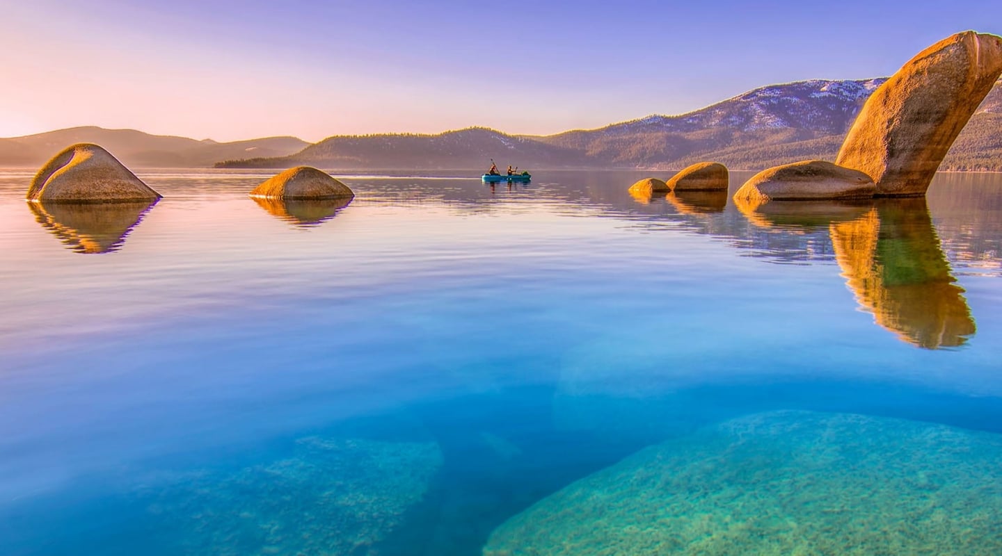 A serene lake scene with kayakers and boulders in the water, surrounded by mountains.