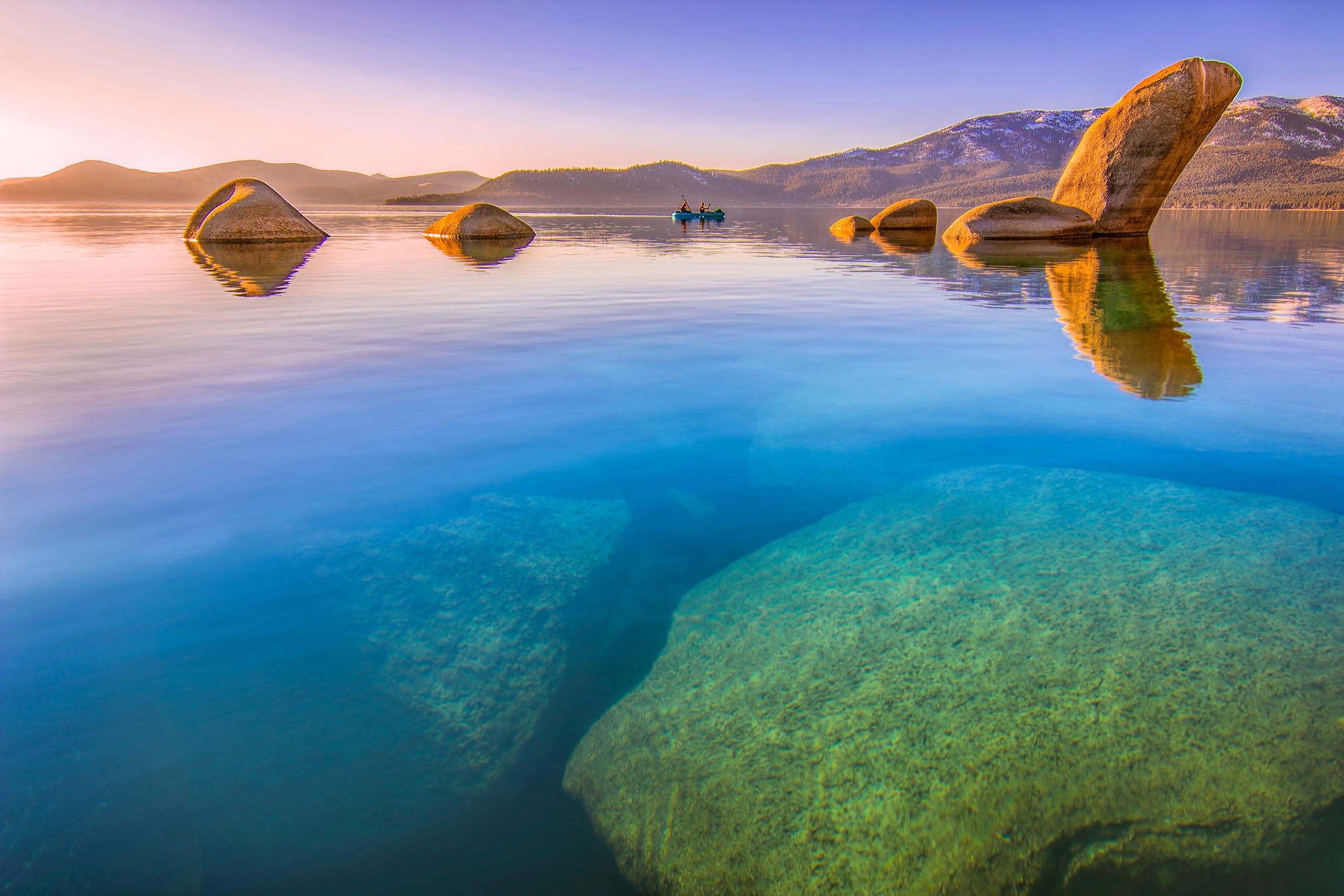 A serene lake scene with large rocks, a boat, and mountains in the background.
