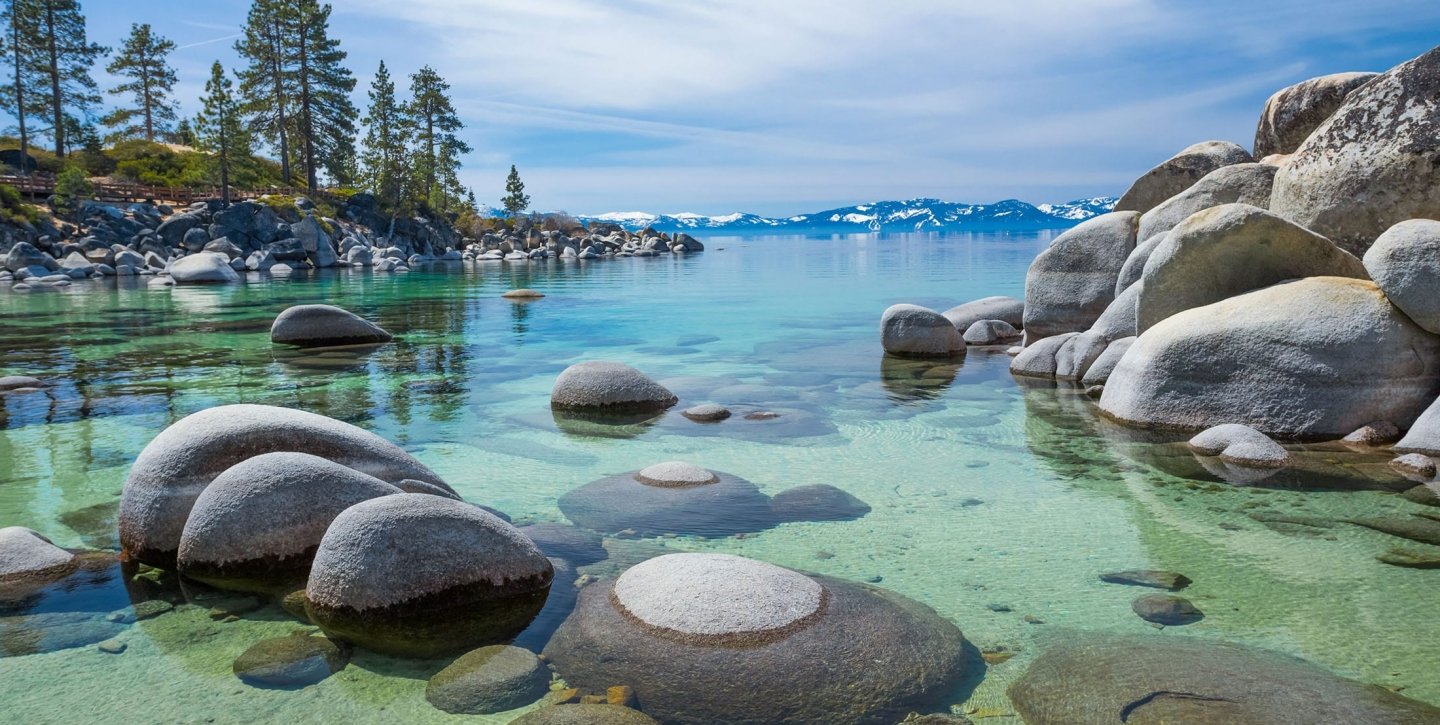Rock formation on the water next to pine trees, mountains and a clear blue sky