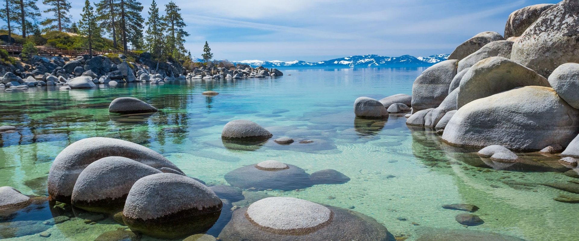 Rock formation on the water next to pine trees, mountains and a clear blue sky