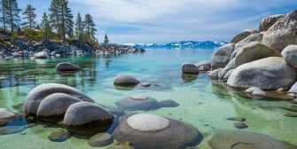 Rock formation on the water next to pine trees, mountains and a clear blue sky