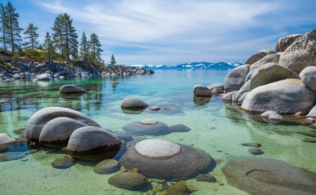 Rock formation on the water next to pine trees, mountains and a clear blue sky