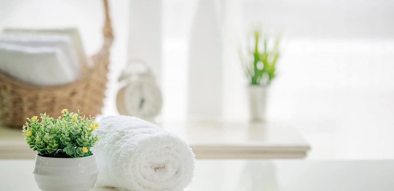 A rolled white towel and a potted plant on a table with a wicker basket and clock in the background.