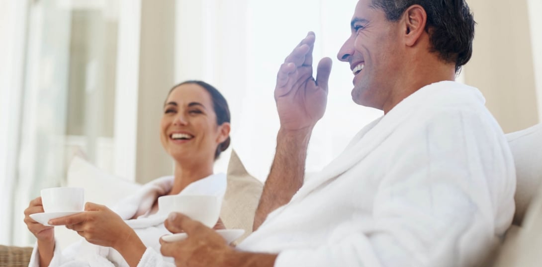 Two people in white robes sitting on a sofa with coffee cups