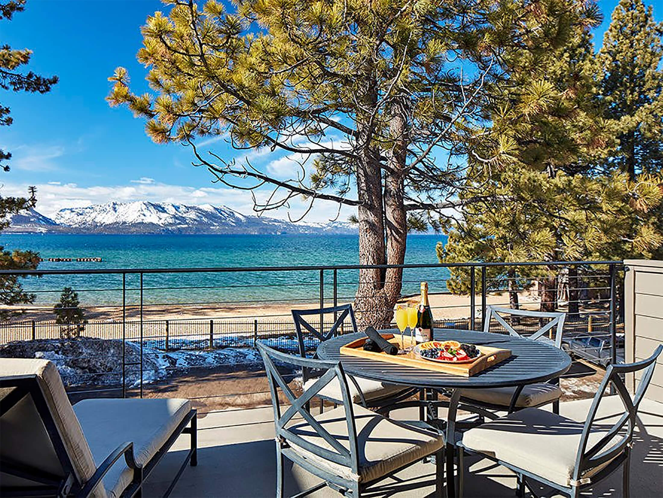 A scenic outdoor dining area with a table set for wine and charcuterie, overlooking a lake with snow-capped mountains in the background.