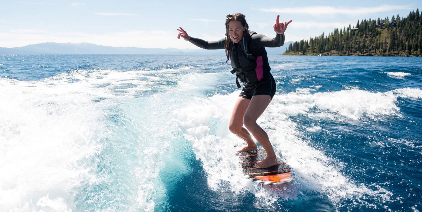 A woman surfing in the blue waters beside the tall pine trees