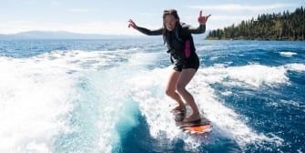 A woman surfing in the blue waters beside the tall pine trees