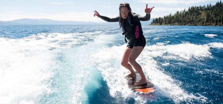 A woman surfing in the blue waters beside the tall pine trees