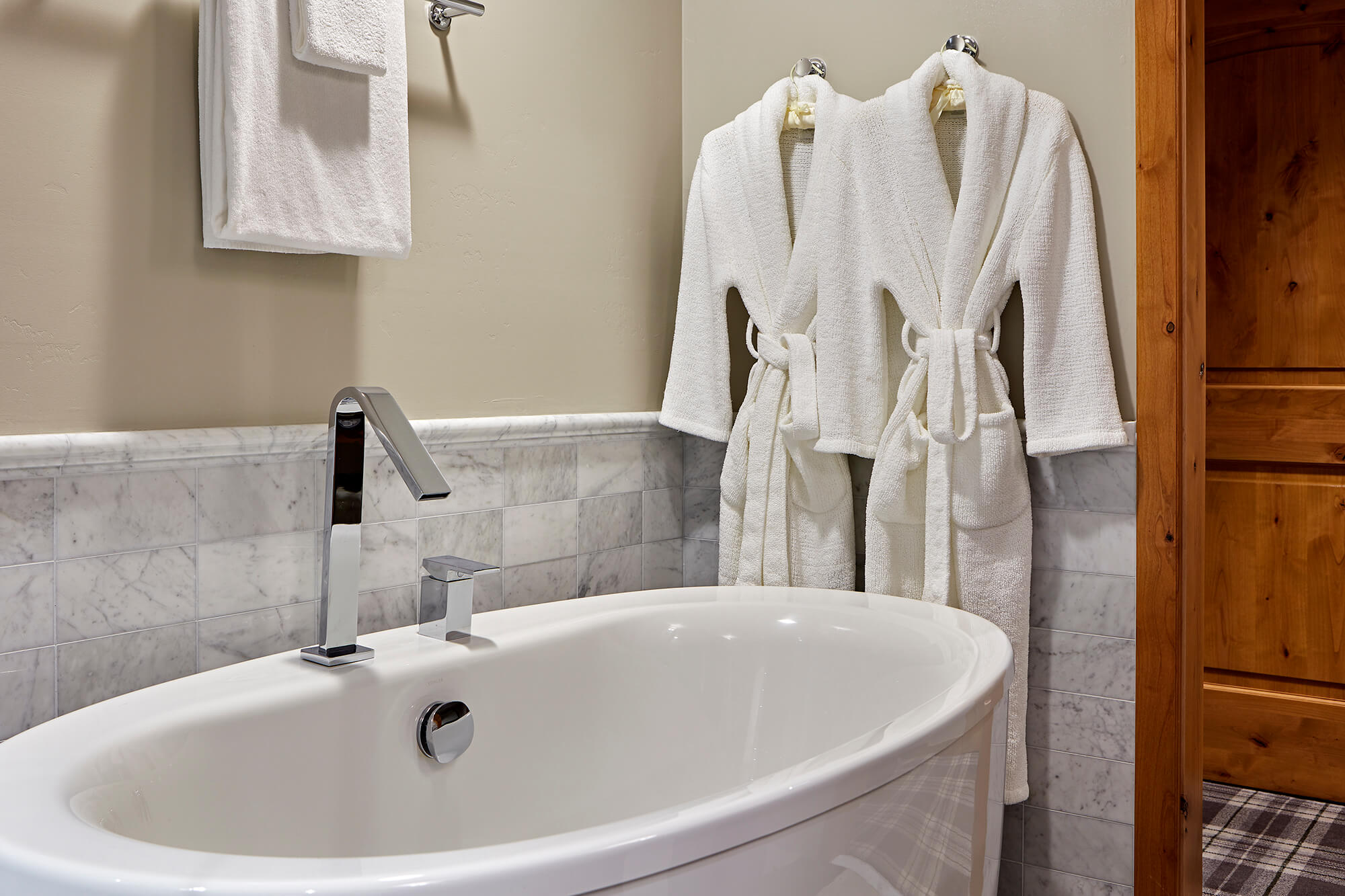 A white soaking tub with a silver faucet, marble tile surround, and two white robes hanging on hooks