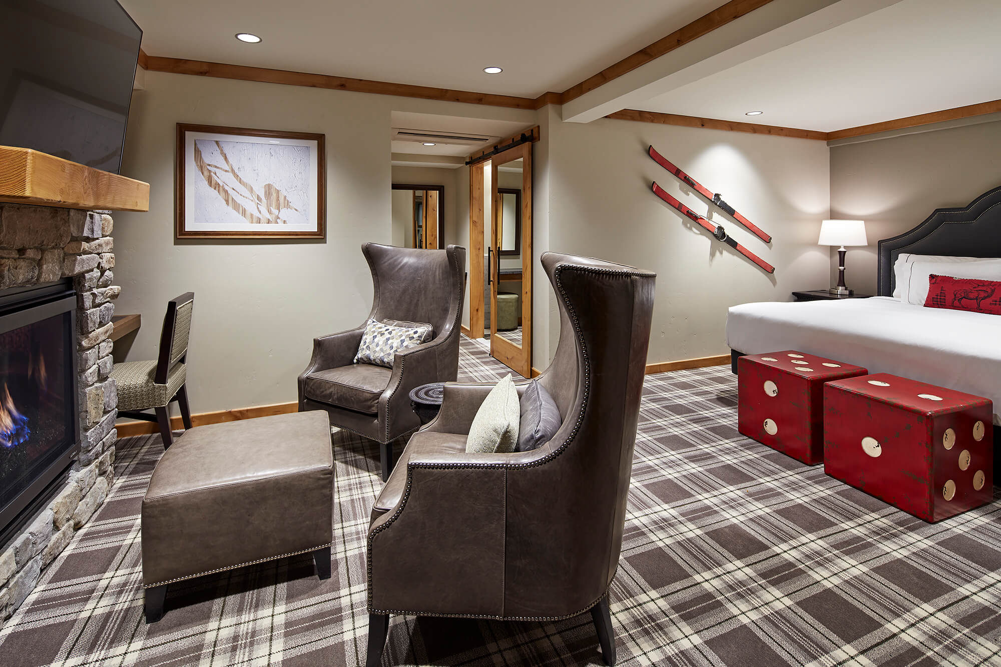 A hotel room with a plaid carpet, stone fireplace, two wingback chairs, a bed with a red pillow, and ski decor on the wall.