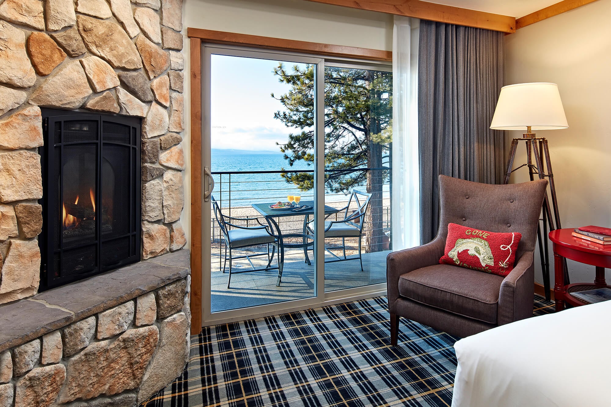 A room with a stone fireplace, plaid carpet, and a sliding glass door leading to a balcony with a view of the water.