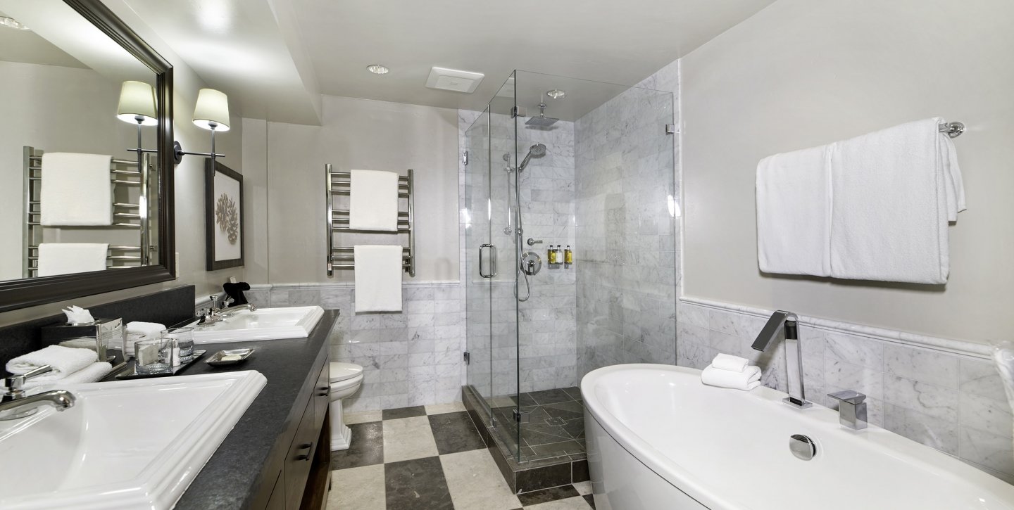 Wooden bathroom countertop with twin marble sinks next to a wide mirror facing the bathtub and shower stall