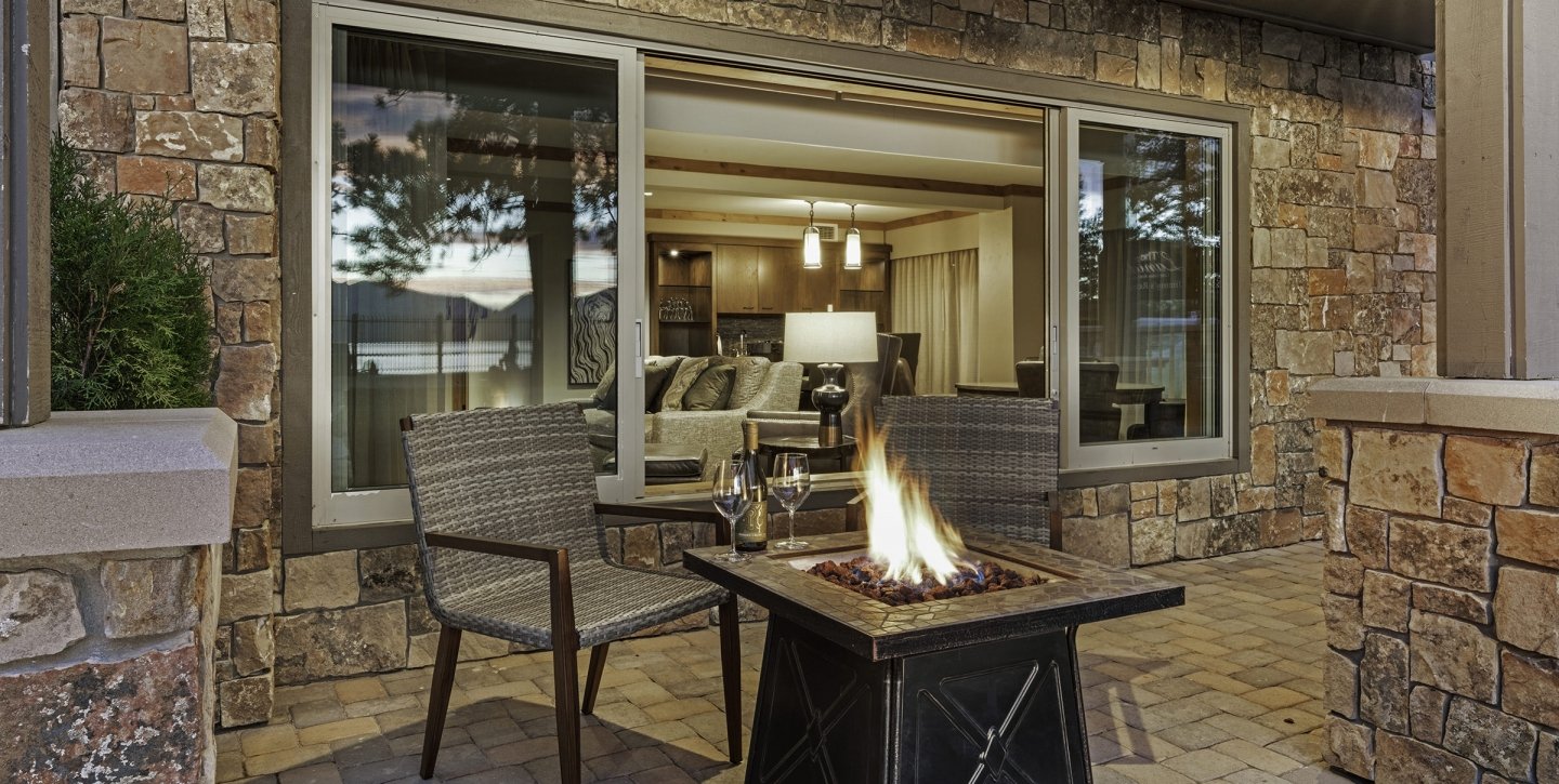 A stone wall with a large window behind the patio with a fire-pit and chairs