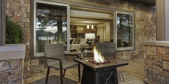 A stone wall with a large window behind the patio with a fire-pit and chairs