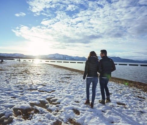A rear view of a man and woman walking in the snow-covered ground next to the lake and mountains
