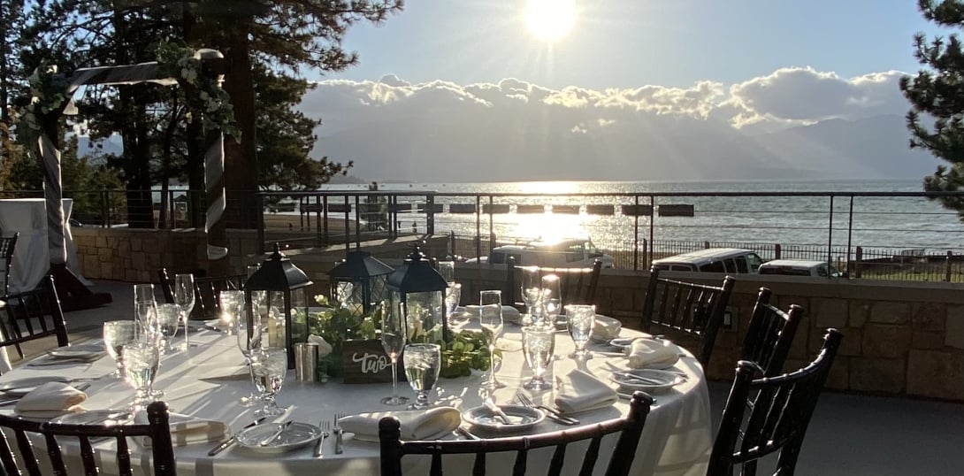 A round dining table set with white tablecloth, wine glasses, and flowers in a outdoor setting with a view of the water and mountains.