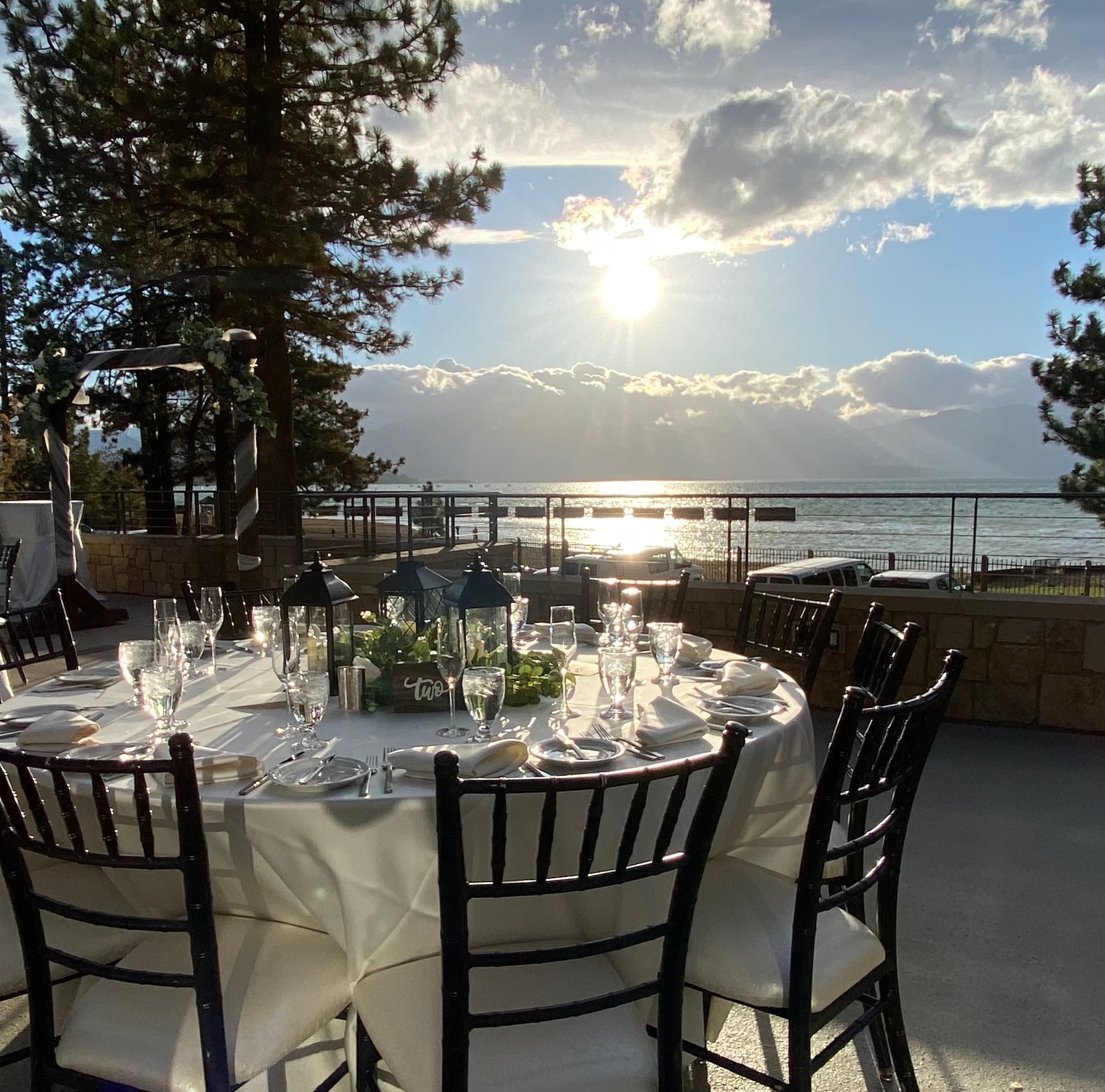 A round dining table set with white tablecloth, wine glasses, and flowers in a outdoor setting with a view of the water and mountains.