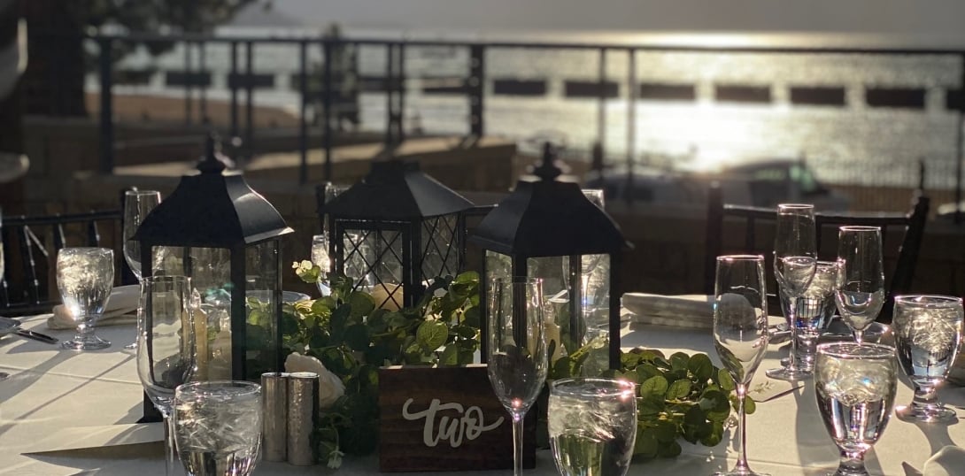 A table set for dining with glasses, lanterns, and greenery on a waterfront patio.