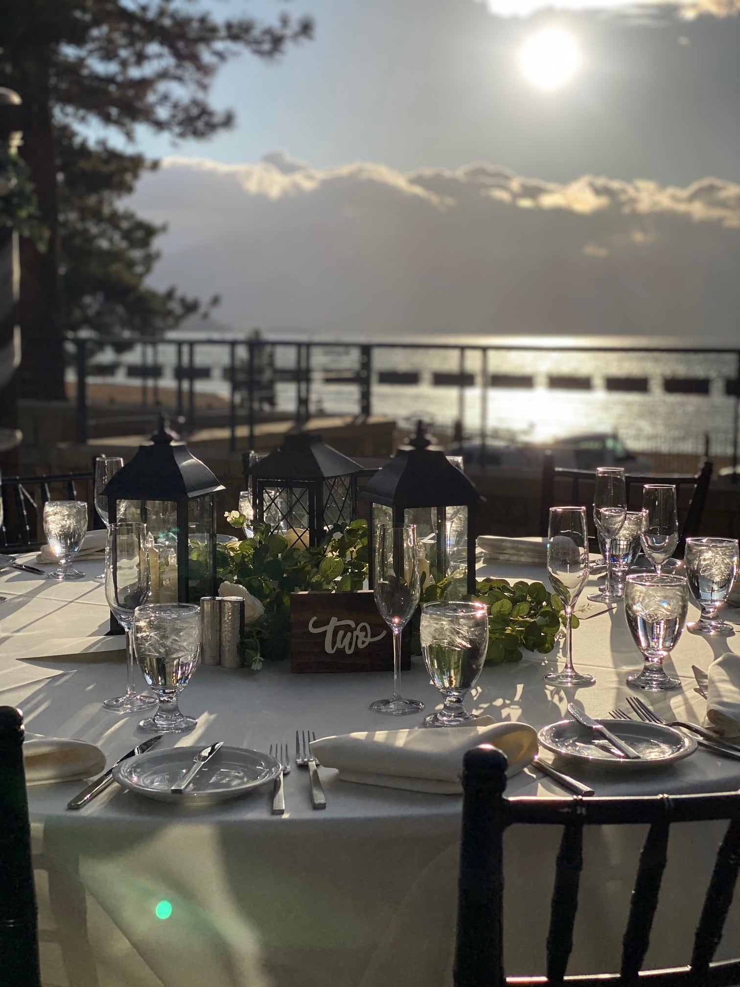 A table set for dining with glasses, lanterns, and greenery on a waterfront patio.
