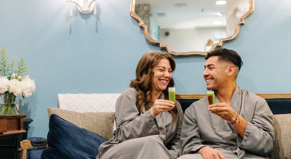 A man and woman in gray robes sit on a couch in a spa lounge.