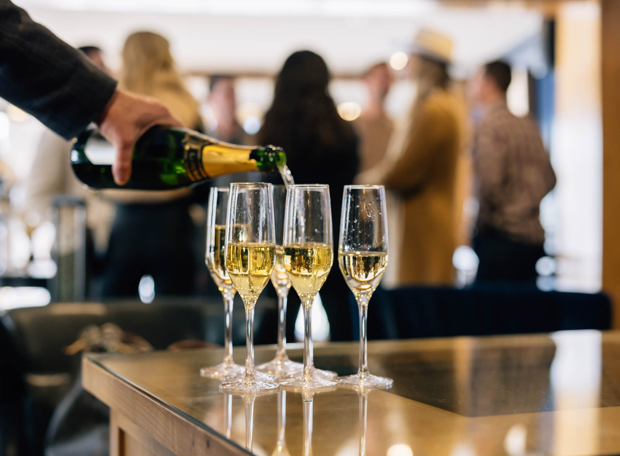A hand pours champagne into four wine glasses on a bar with people in the background.
