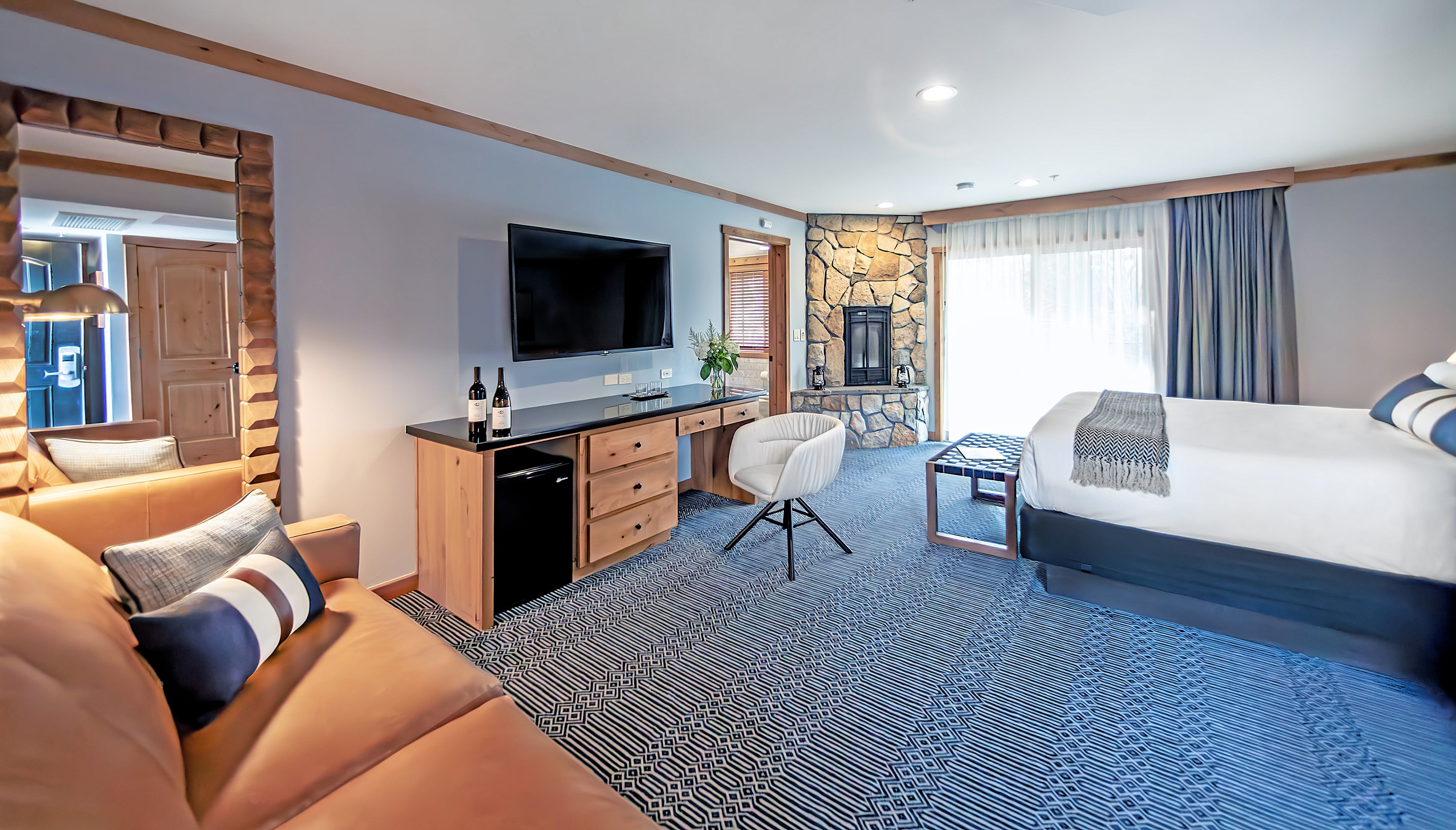 A wide-angle view of a hotel room with a bed, sofa, desk, TV, and fireplace.