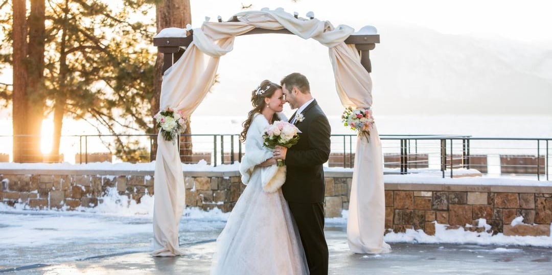 a couple in wedding attire standing under an archway with a stone wall and trees in the background