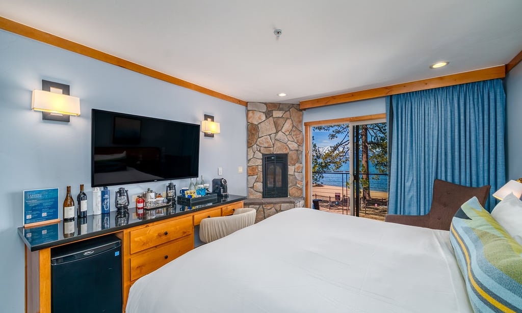 A hotel room with a large bed, stone fireplace, flat-screen TV, and balcony with a lake view.