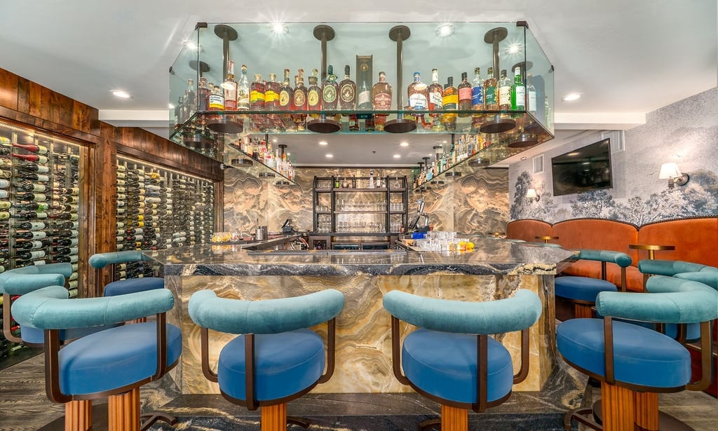A well-stocked bar with a marble countertop, blue stools, and extensive wine and liquor storage.
