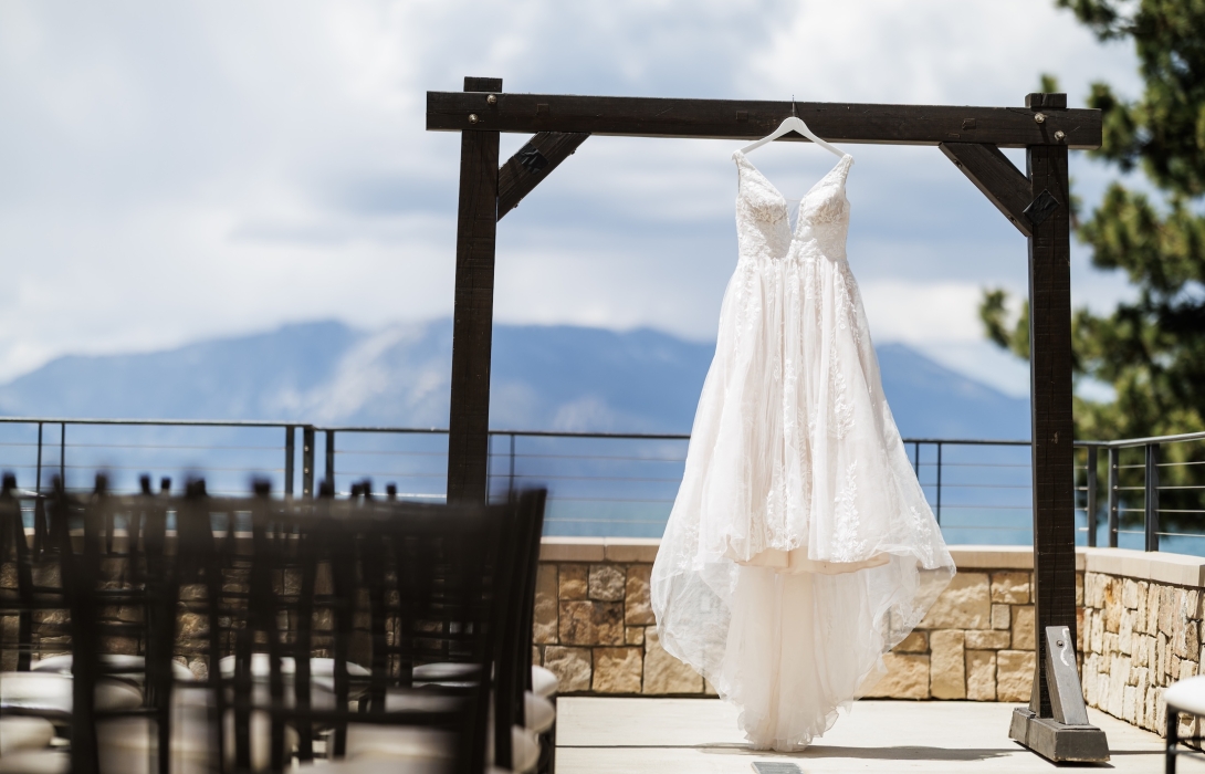 White lace wedding dress hanging on a wooden frame outdoors with a cloudy blue sky and distant mountains behind it