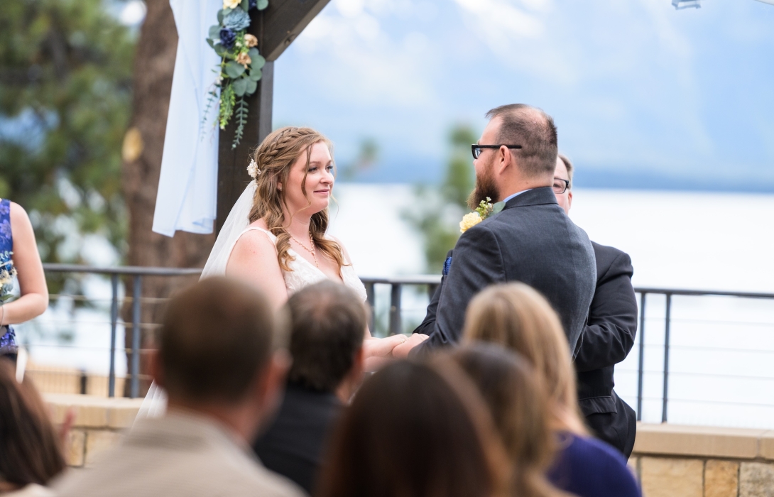 Bride and groom hold hands during an outdoor wedding ceremony beneath a floral arch, with guests seated in the foreground by a railing and lake in the background.
