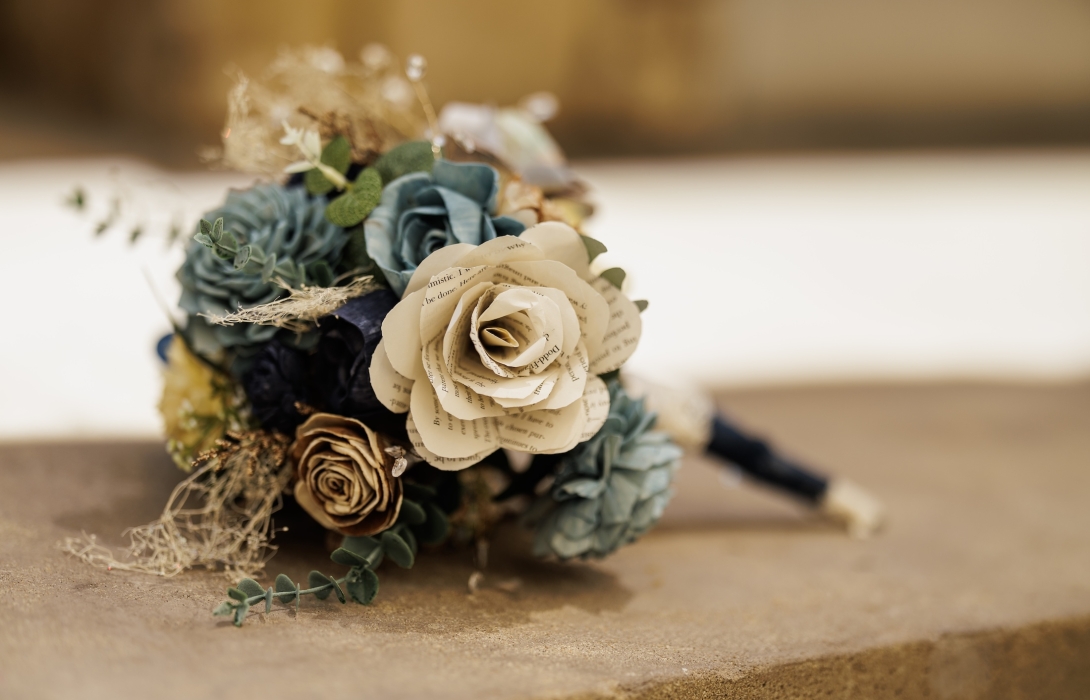 Close-up of a paper flower bouquet with newspaper-printed petals and green foliage resting on a wooden surface.