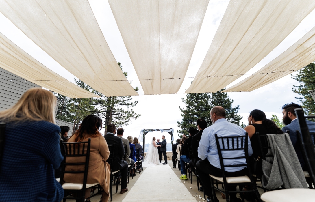 Outdoor wedding ceremony under cream fabric canopies with guests seated on both sides of a white aisle, couple at the altar exchanging vows.