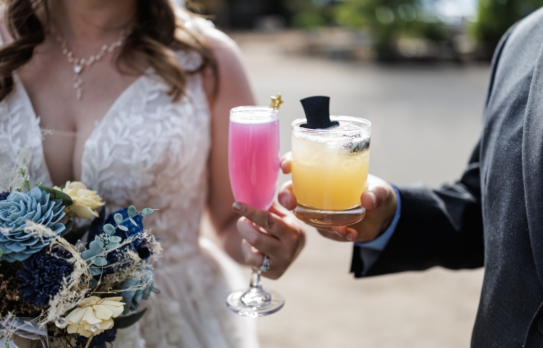 Bride in a lace wedding dress toasting with two colorful drinks (pink and yellow) as a bouquet is visible in the foreground.