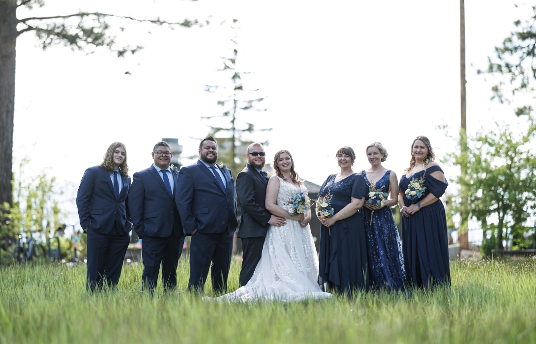 Wedding party posing on a grassy lawn: bride in a white gown, bridesmaids in navy blue dresses, and groomsmen in dark suits smiling together outdoors.