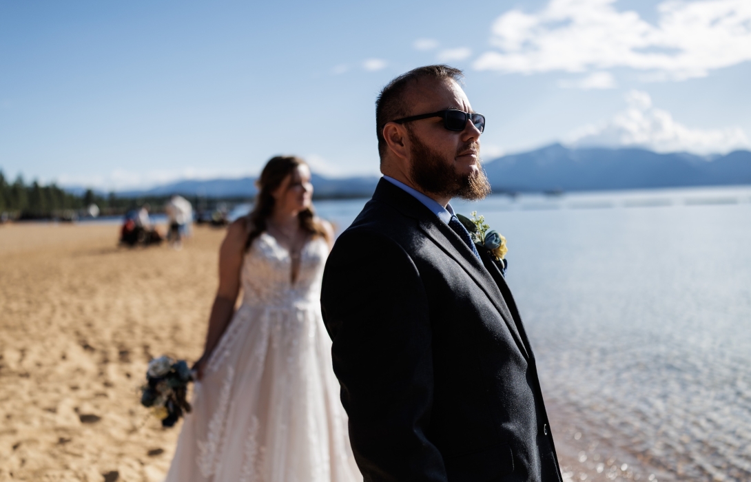 Man in a dark suit and sunglasses stands on a sandy beach with a bride in a white dress blurred in the background.