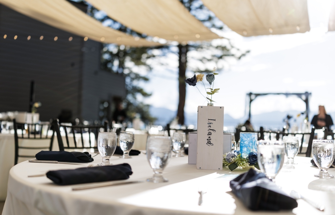Outdoor event table set for a formal occasion with white tablecloths, black napkins, glassware, and a centerpiece sign with dried flowers under string lights during daytime.