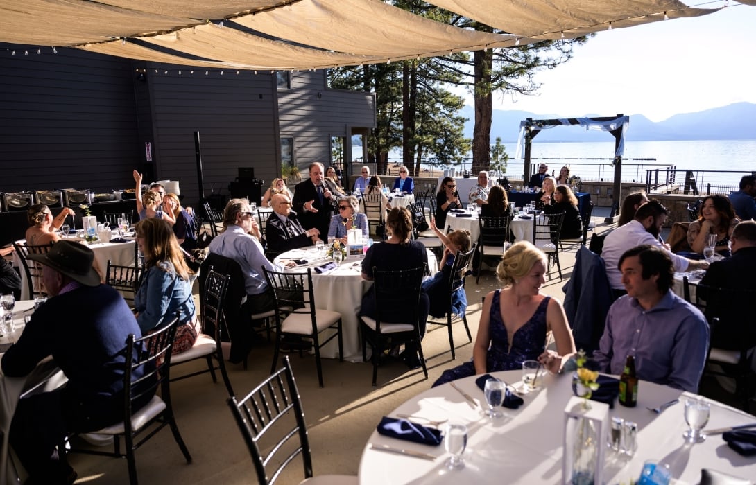 Outdoor lakeside wedding reception with guests seated at round tables under a shade canopy while a man in a suit gives a microphone toast nearby.