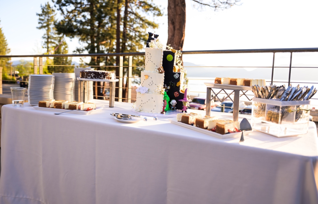 Outdoor dessert table on a patio with a tall white cake, plates, and assorted pastries; railing, trees, and lake in the background