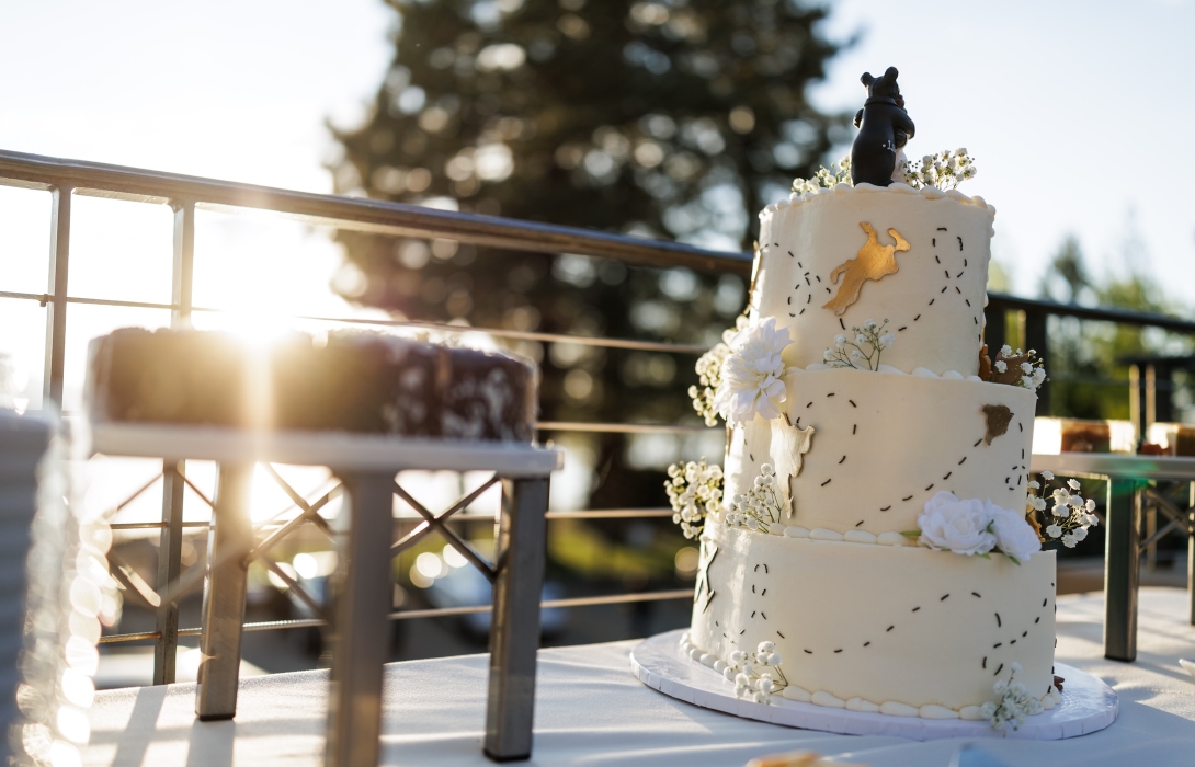 Three-tier white wedding cake with a black dog figurine topper, white flowers, and gold map accents in a sunny outdoor setting.