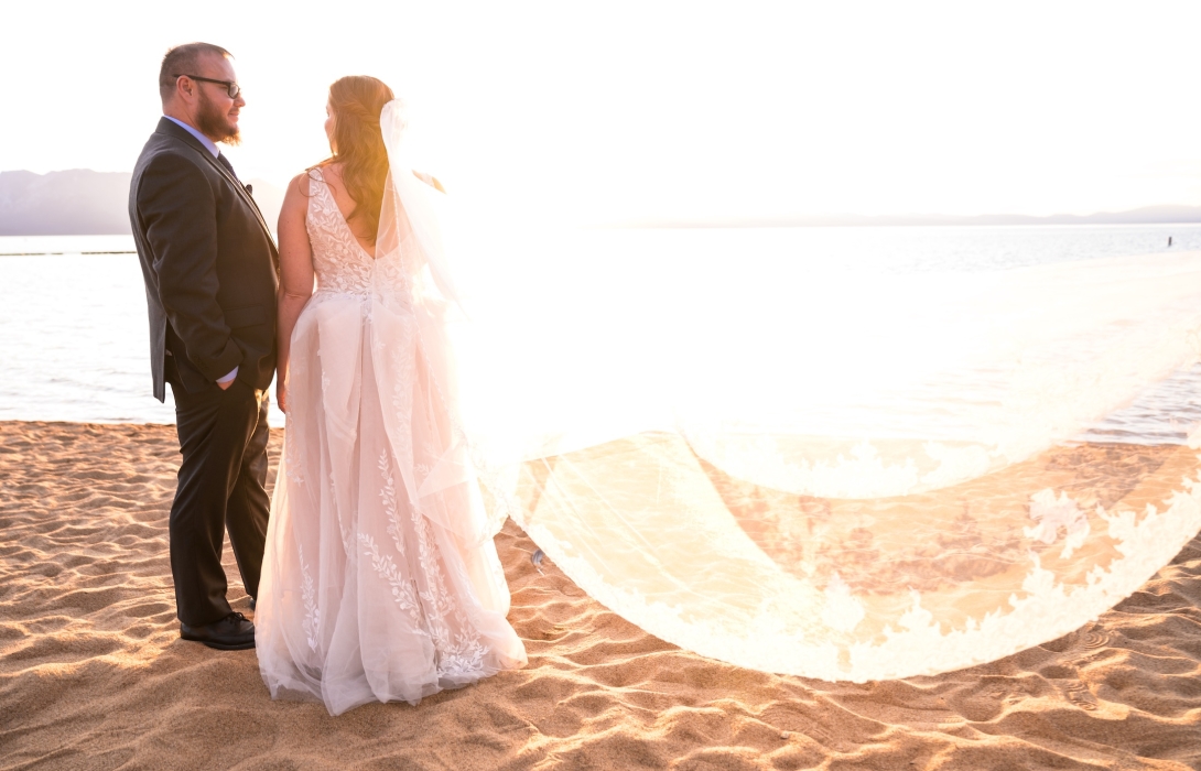 Bride and groom stand on a sandy beach at sunset, facing each other in wedding attire by the water.