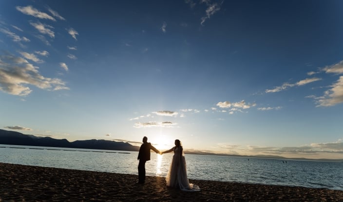 Couple on a sandy beach holding hands at sunset with the sun low over calm water and mountains in the distance