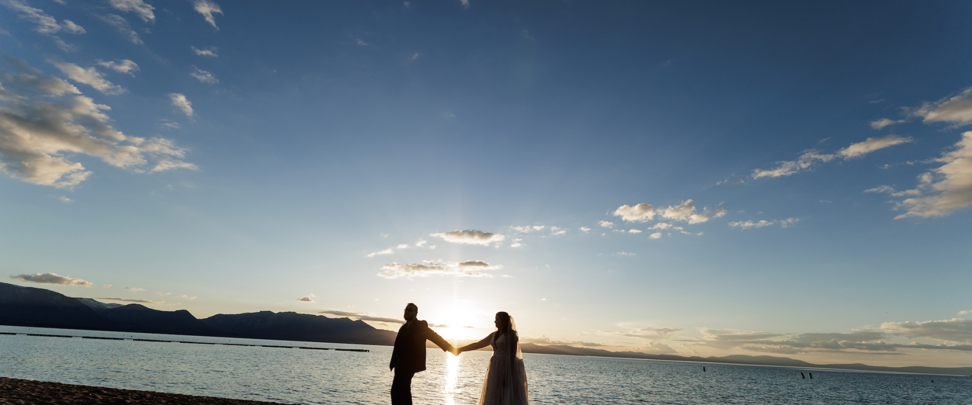 Silhouetted couple on a sandy beach at sunset, holding hands with the sun low over calm water.