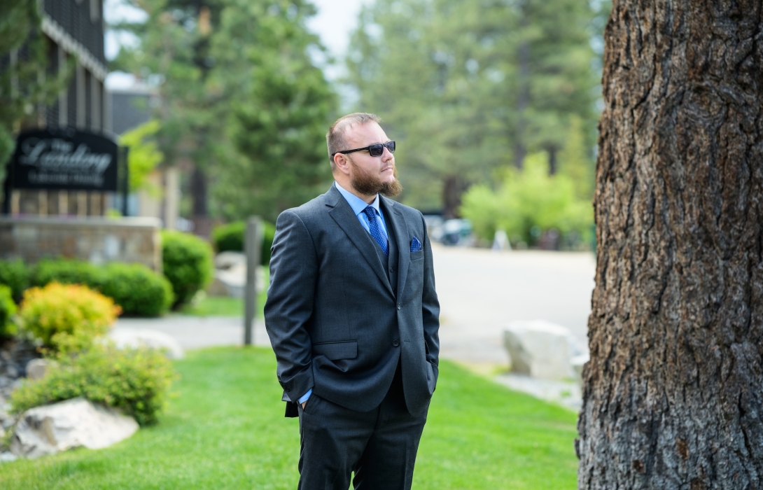 Man in a blue suit and sunglasses stands outdoors beside a large tree, looking off to the distance.