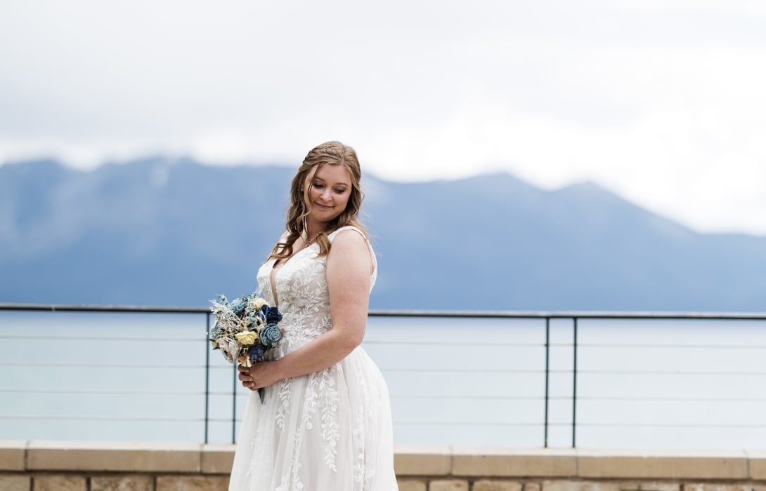 Bride in a white lace wedding gown holding a bouquet, standing on a terrace with a stone wall and distant mountains.