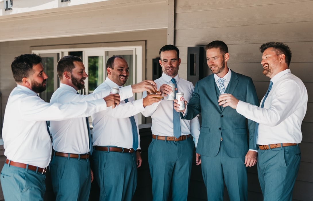 Six groomsmen in teal pants and white shirts toast with cans and glasses on a wooden porch.