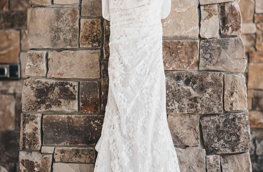 White lace wedding dress hanging on a white hanger against a textured stone wall.